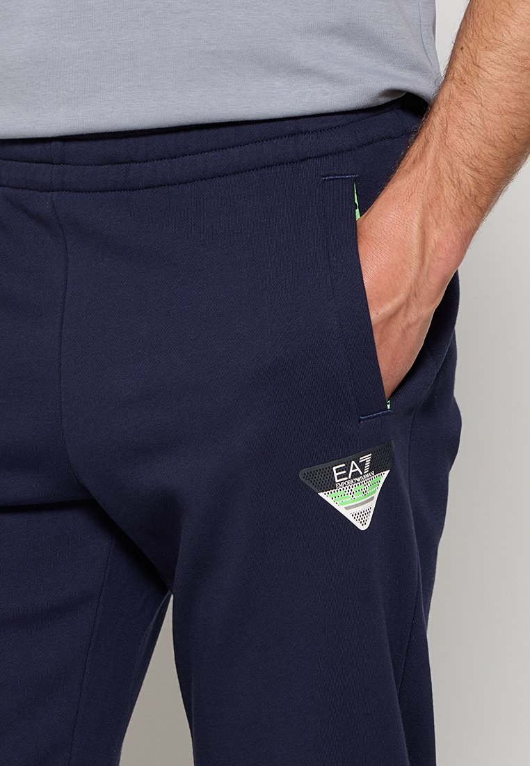 Granatowe dresy wykonane z miękkiego materiału, z boczną kieszenią i naszywką logo EA7 w kolorze czarnym i zielonym, z trójkątnym wzorem.