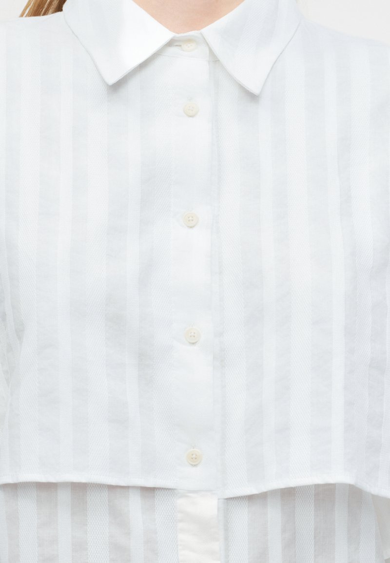 Chemise blanche à col avec des rayures verticales transparentes et quatre boutons visibles sur le devant, portée par une personne dont le cou est tronqué.