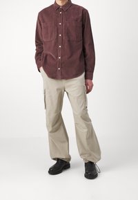 Camisa de pana marrón con botones y dos bolsillos frontales, combinada con pantalones cargo beige y botas negras hasta el tobillo. Fondo liso.