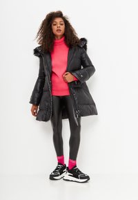Cappotto a pannello nero con cappuccio, caratterizzato da un bordo in pelliccia sintetica, dettagli in argento e una texture lucida. Indossato sopra un maglione rosa e leggings neri.