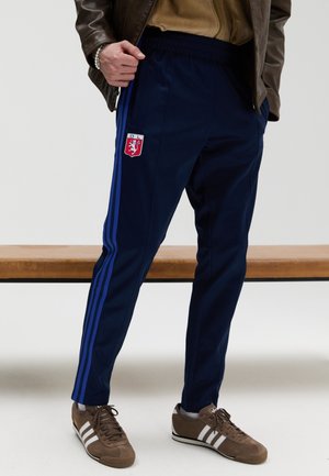 Osoba nosząca granatowe spodnie dresowe z niebieskimi paskami i czerwonym herbem klubu Lyon, w połączeniu z brązowymi butami Adidas i brązową kurtką.