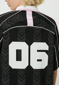 Person med kort blont hår som bär en svart sporttröja med vit nummer 06, rosa och vit krage samt chevronmönster på ryggen.