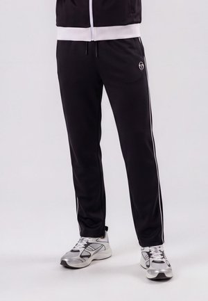 Partie inférieure du corps portant un pantalon de survêtement noir avec des bandes blanches sur les côtés et des chaussures de sport blanches et grises sur un fond uni.