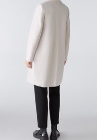 Abrigo de lana gris claro con un acabado texturizado, diseño entallado y corte de longitud media. Combinado con pantalones negros y zapatos de charol.