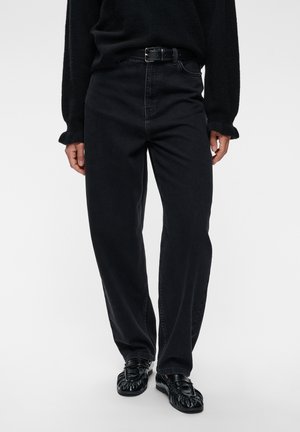 Personne portant un jean noir taille haute avec une ceinture noire, un pull noir et des chaussures en cuir noir debout sur un fond blanc.
