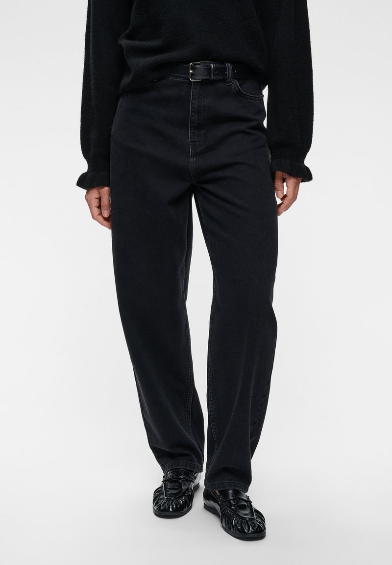 Personne portant un jean noir taille haute avec une ceinture noire, un pull noir et des chaussures en cuir noir debout sur un fond blanc.
