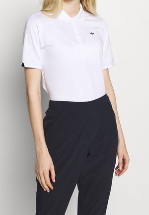 Vrouw draagt een wit polo met korte mouwen met Lacoste-logo en hoge zwarte broek, staand tegen een effen achtergrond.