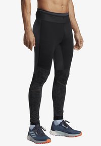 Black athletic leggings with a fitted design, featuring textured accents and a zippered pocket. Pair of blue and orange sneakers completes the look.