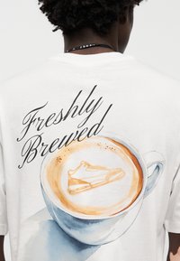 Λευκό βαμβακερό t-shirt με υδατογραφία ενός φλιτζανιού καφέ, που έχει σχεδιασμό αθλητικού παπουτσιού στον αφρό και το κείμενο "Φρεσκοκαβουρδισμένος".