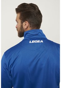 Giacca sportiva blu con colletto alto e tessuto testurizzato. Logo "LEGEA" in bianco sulla schiena, con un design aerodinamico.