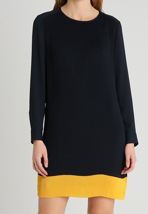 Femme portant une robe noire à manches longues avec un ourlet jaune contrastant, debout devant un fond clair uni.