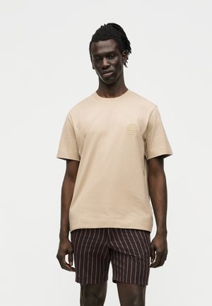 Jeune homme à la peau foncée portant une chemise beige à manches courtes et un short marron foncé à fines rayures, debout sur fond blanc.