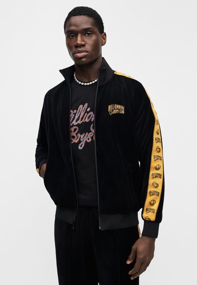 Μαύρο βελούδινο τζάκετ φόρμας με κίτρινες ρίγες στο πλάι, με το λογότυπο "Billionaire Boys Club". Γιακάς που στέκεται και κλείνει με φερμουάρ.