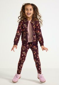 Marnblomstret træningsdragt med lynlåsjakke og matchende leggings, der har farverige blomstermønstre. Lys rosa top og pink sneakers.