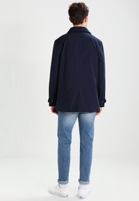 Marineblå letvægtsfrakke med klassisk krave, slids i ryggen og knappede manchetter, kombineret med lyseblå denimjeans og hvide sneakers.