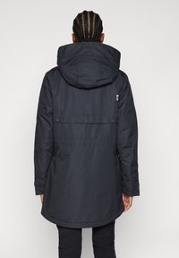 Person med flätat hår som bär en lång svart huva jacka, stående med ryggen mot en slät ljus bakgrund.