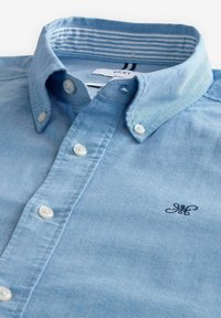 Camisa de botones azul claro hecha de algodón. Presenta un cuello con solapa, botones blancos y un logo bordado en el lado izquierdo del pecho.