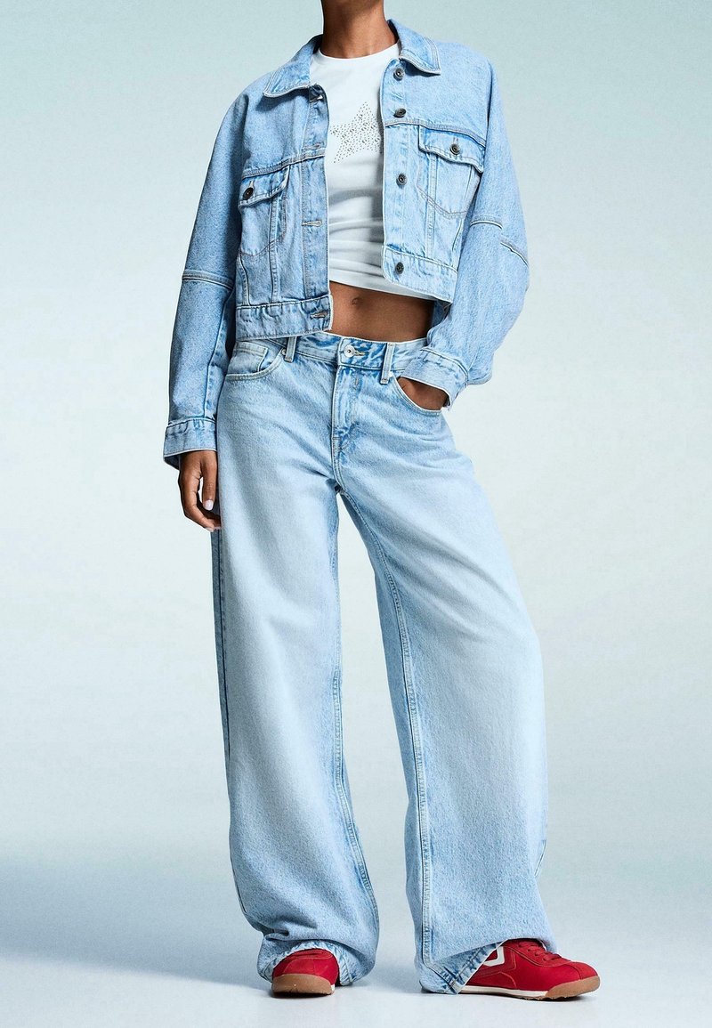 Ljusblå jeansjacka med kort design och guldfärgade detaljer, kombinerad med vida ljusblå jeans. Röda sneakers fulländar outfiten.