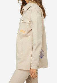 Veste beige texturée avec deux poches avant. Ornée d'un symbole infini orange et d'une broderie de coquillage violette. Dotée d'un col avec un revers.