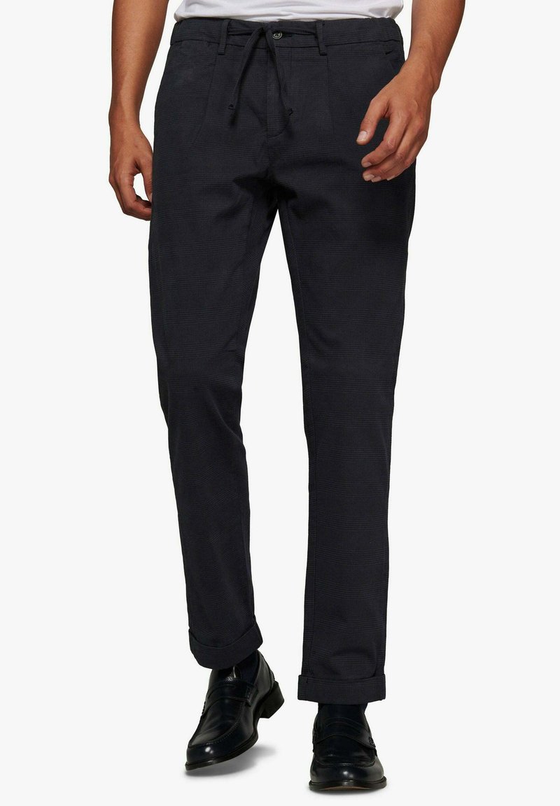 Pantalones oscuros de color marino con una tela texturizada, que presentan una cintura con cordón, bolsillos laterales y puños enrollados. Combinados con zapatos negros.