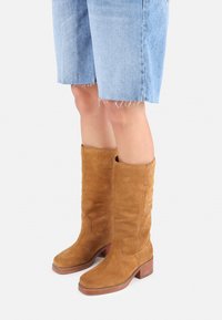 Tan suede knee-high boots with a block heel and stitched details, paired with frayed denim shorts, shown against a neutral background.