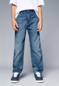 Zila džinsu bikses ar brīvu piegriezumu, kas raksturojas ar tradicionālo piecu kabatu dizainu un vidēju jostas vietu, valkātas kopā ar tumši ziliem sporta apaviem un baltu krekliņu.