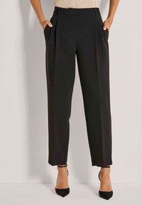 Vrouw in een hooggewaist zwarte pantalon met zijzakken en zwarte puntige enkelsandalen met enkelbandje, staand tegen een lichte muur.