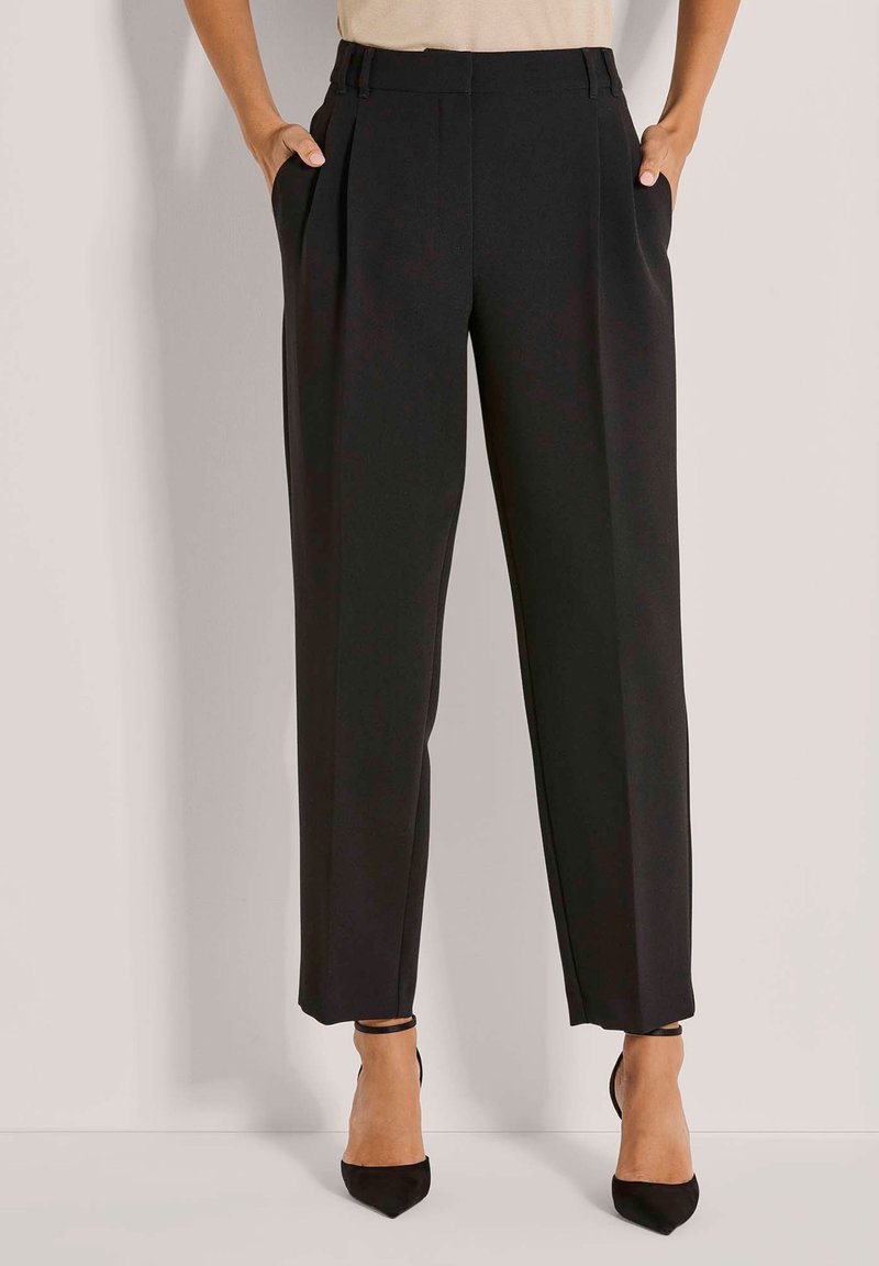 Vrouw in een hooggewaist zwarte pantalon met zijzakken en zwarte puntige enkelsandalen met enkelbandje, staand tegen een lichte muur.