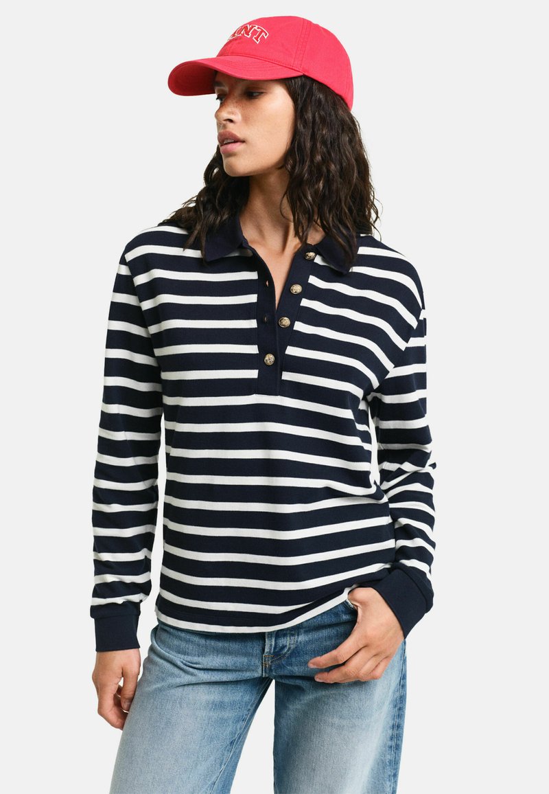 Marineblauw en witte gestreepte polo met lange mouwen, kraag en een drieknoopsluiting. Gecombineerd met lichtblauwe jeans en een roze honkbalpet.