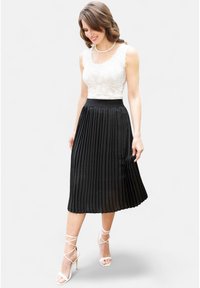 Gonna nera plissettata con una texture morbida, abbinata a un top in pizzo bianco. La modella indossa sandali con tacco alto bianchi e con strappi.
