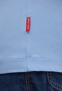 Hellblauer Stoff mit vertikalen und horizontalen Nähten, rotes rechteckiges Etikett mit der Aufschrift "JOOP! JEANS", darüber dunkelblauer Denim mit oranger Naht.