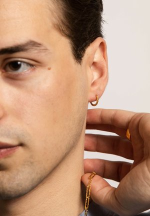 Goldene Creolen mit einem gedrehten Design, die am Ohr getragen werden, kombiniert mit einer zarten goldenen Kette, die zwischen den Fingern gehalten wird und eine klare Textur zeigt.