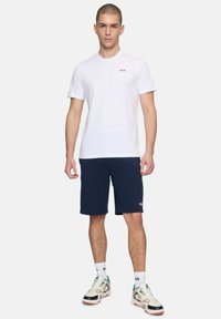 Witte t-shirt met korte mouwen en Fila-logo, gecombineerd met marineblauwe shorts. Model draagt meerkleurige sneakers met contrasterende accenten en witte sokken.