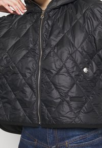 Černá prošívaná bunda se zipem vpředu, kapucí a logem při lemu. Měkký, lesklý materiál s diamantovými vzory a volným střihem.