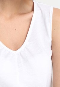 Vit ärmlös topp med V-ringning, tillverkad av lätt tyg med subtil textur och en draperad detalj vid ringningen.