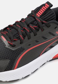 Chaussure de sport noire avec des accents rouges, tige en maille, semelle texturée et lacets noirs. Se distingue par un logo de marque bien en vue et un design épuré.