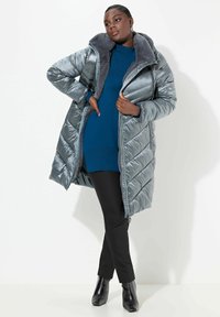 Giubbotto a sfera argento con colletto in pelliccia sintetica, caratterizzato da un motivo a zigzag, indossato sopra un top blu e pantaloni neri, abbinato a stivali neri con punta.