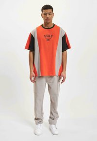 Tricou supradimensionat portocalie și gri cu accente negre și logo, asortat cu pantaloni gri deschis cu cusături laterale și logo. Adidași albi.
