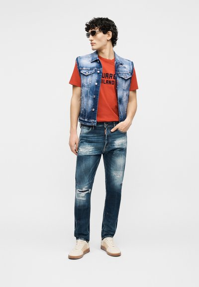 Νεαρός άνδρας που φοράει γυαλιά ηλίου, αμάνικο μπλε τζιν γιλέκο πάνω από κόκκινη μπλούζα με σχέδιο, ξεσκισμένο μπλε τζιν και λευκά αθλητικά παπούτσια, στέκεται με το χέρι στην τσέπη.