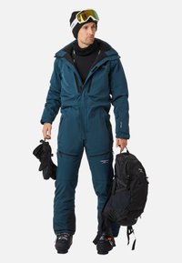Teal weerbestendige skibroek met een gestructureerde pasvorm, met rits afgesloten zakken, zwarte accenten en een zwarte rugzak in één hand vastgehouden.