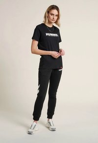 Melnā T-krekls ar baltu "HUMMEL" logotipu, kombinēts ar melnām sporta biksēm ar baltām ķiveres detaļām. Sporta apavi ar pelēkām un baltām detaļām.