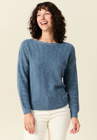 Maglione blu lavorato a maglia con un motivo a spina di pesce testurizzato, scollatura ampia e maniche lunghe. Indossato con pantaloni chiari.