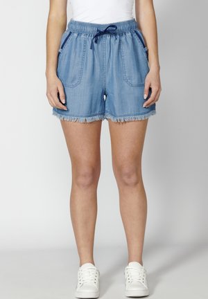 Denim kratke hlače s elastičnim strukom, detaljem za vezanje i prednjim džepovima. Imaju oštri rub i svijetlu plavu boju. Uparene su s bijelim tenisicama.