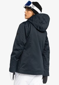 Marineblauwe geïsoleerde jack met capuchon, uitgerust met een gladde textuur, verstelbare manchetten en een logo patch op de mouw. Witte handschoenen zichtbaar.