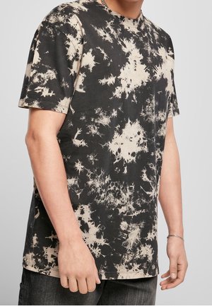 Mężczyzna ubrany w czarno-beżowy t-shirt z krótkim rękawem w stylu tie-dye oraz ciemne spodnie, widoczny od ramion do połowy uda na jednolitym tle.