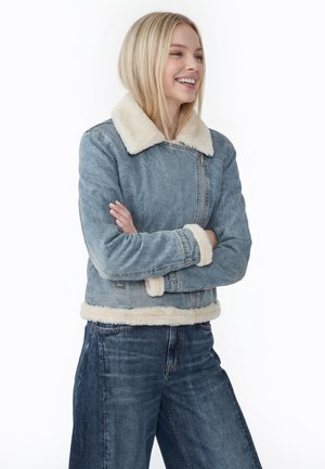Lachende vrouw met blond haar die een lichtblauwe spijkerjas met witte bontafwerking en bijpassende wijde spijkerbroek draagt.