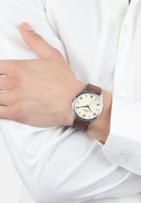 Brauner Lederarmbanduhr mit silbernem Gehäuse, cremigem Zifferblatt, auffälligen schwarzen Stundenmarkierungen und einem Datumsfenster auf der 3-Uhr-Position, getragen an einer Hand.