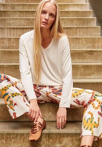 Witte lange mouwen top met een V-hals, gestreepte wijde broek met oranje en blauwe dierenpatronen, en bruine veterschoenen.