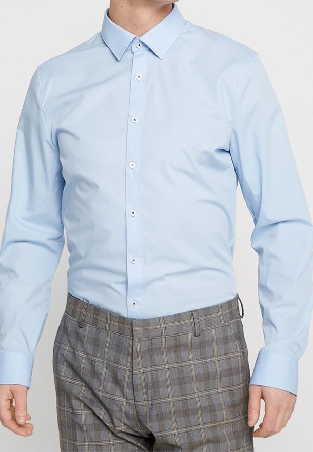 Homme portant une chemise habillée bleu clair boutonnée et un pantalon gris à carreaux, debout les bras détendus le long du corps.