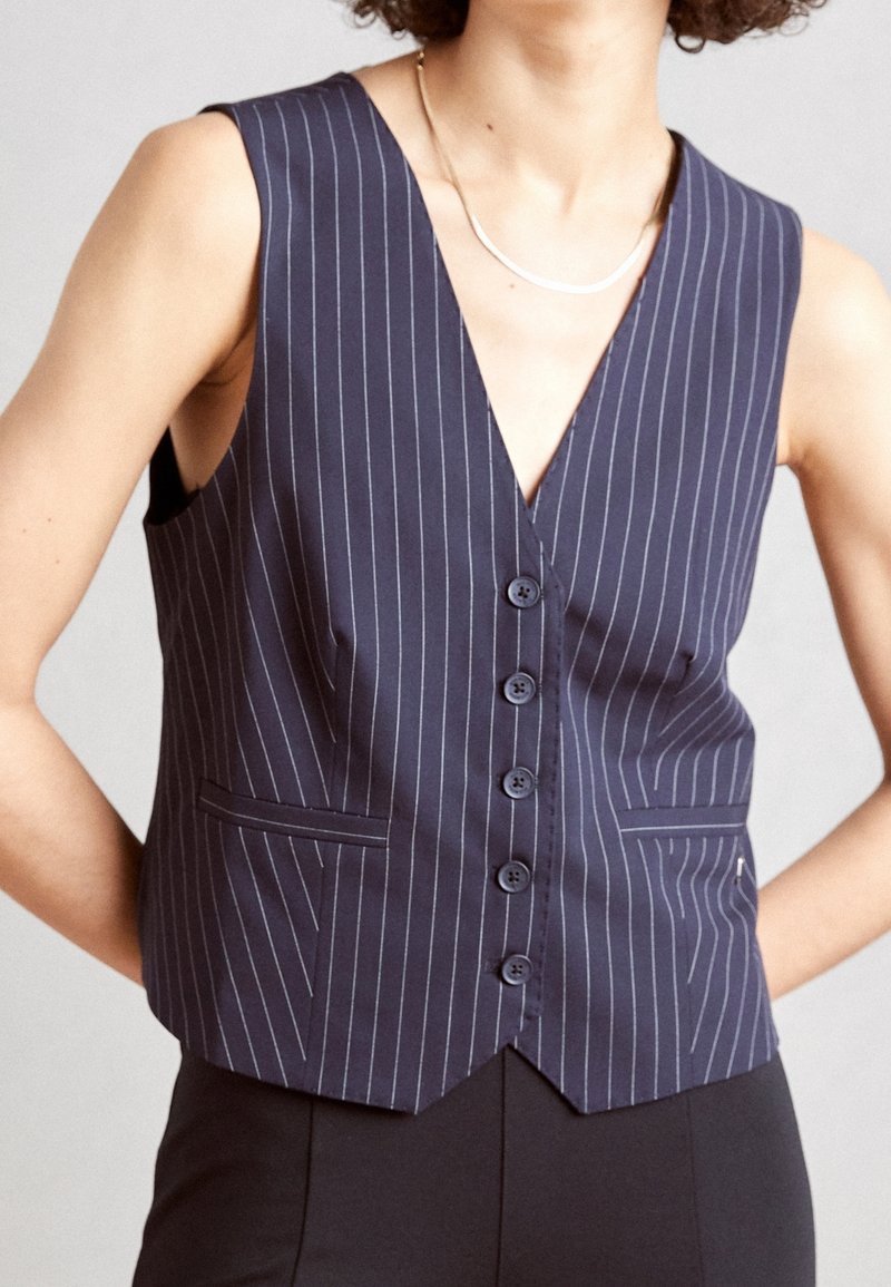 Marinblå väst med smalrand, djupt V-ringad hals, fem framknappar och två sidofickor. Tillverkad av ett mjukt tyg med en skräddarsydd passform.